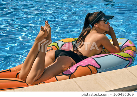 Portrait of a female having rest and posing in a swimming pool on an inflatable mattress. Dressed in a black swimsuit, sun visor and sunglasses. Portrait of a female having rest and posing in a swimming pool on an inflatable mattress. Dressed in a black swimsuit, sun visor and sunglasses. 100495369