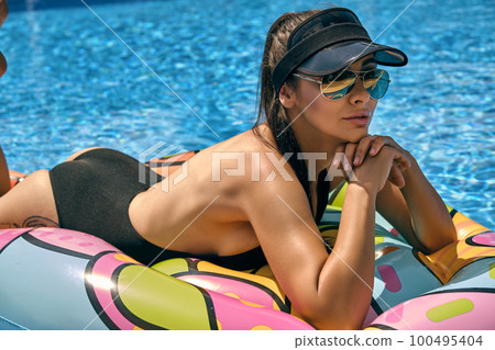 Portrait of a female having rest and posing in a swimming pool on an inflatable mattress. Dressed in a black swimsuit, sun visor and sunglasses. 100495404