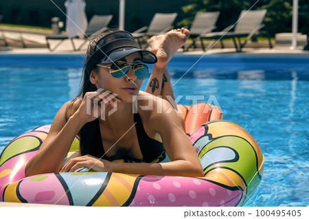 Portrait of a female having rest and posing in a swimming pool on an inflatable mattress. Dressed in a black swimsuit, sunvisor cap and sunglasses. 100495405