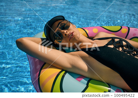 Portrait of a female having rest and posing in a swimming pool on an inflatable mattress. Dressed in a black swimsuit, sun visor and sunglasses. Portrait of a female having rest and posing in a swimming pool on an inflatable mattress. Dressed in a black swimsuit, sun visor and sunglasses. 100495422