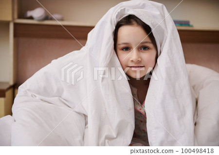 Adorable Caucasian little child girl hiding in white soft blanket, looking at camera, cutely smiling looking at camera. The concept of childhood, daily routines and bedtime 100498418