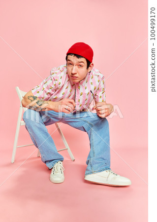Stylish guy grimacing and leaning forward and posing while sitting on chair over pink background. Concept of human emotions, fashion, youth, student life 100499170