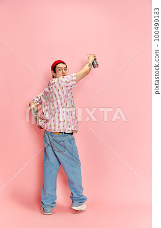 Tattooed graffiti urban painter with spray starting to draw graffiti on the wall over pink background. Full length portrait. Copy space for ad Tattooed graffiti urban painter with spray starting to draw graffiti on the wall over pink background. Full length portrait. Copy space for ad 100499183