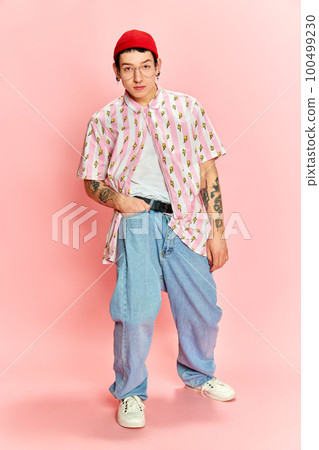 Portrait of stylish guy with tattoo keeping hand in pocket and looking at camera over pink background. 100499230