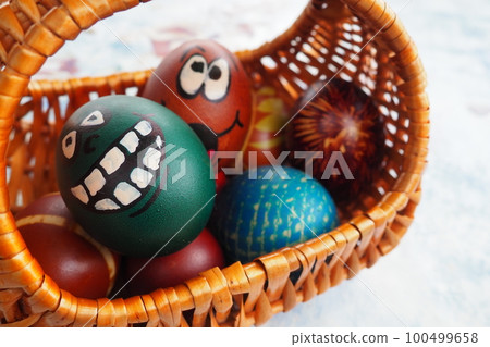 Easter eggs painted in different colors with painted laughing faces. Funny grimaces with eyes, tongue and big white teeth. Scary face for Halloween. Emoticon for Easter Wicker basket. White background 100499658
