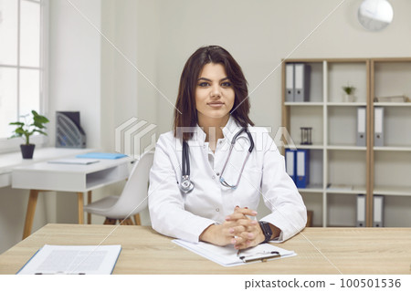Portrait of young confident female doctor or nurse sitting at desk at her workplace. Portrait of young confident female doctor or nurse sitting at desk at her workplace. 100501536