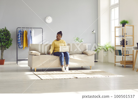 Young African American woman working on her laptop while sitting on the sofa at home Young African American woman working on her laptop while sitting on the sofa at home 100501537