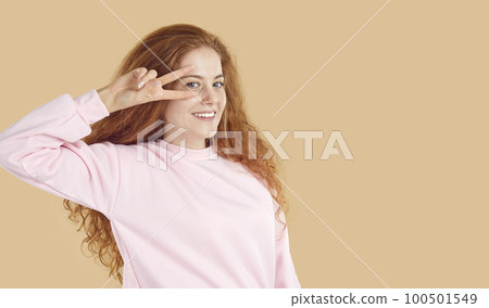 Portrait of cute Caucasian teenage girl making peace gesture showing v-sign near eye. 100501549
