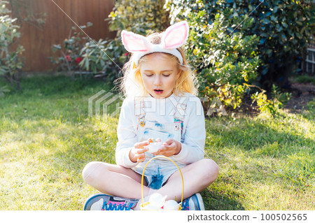 Little girl wearing bunny ears holding opened container with Easter chocolate egg sweets,candies after egg hunting in garden. Kid having fun on Easter holiday. Children eating sweets. Selective focus Little girl wearing bunny ears holding opened container with Easter chocolate egg sweets,candies after egg hunting in garden. Kid having fun on Easter holiday. Children eating sweets. Selective focus 100502565