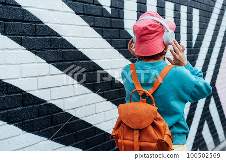 Back view stylish woman in bright clothes and bucket hat wearing wireless headphones and listening to music while walking near city urban painted wall. Fashionable hipster lifestyle. Selective focus. 100502569