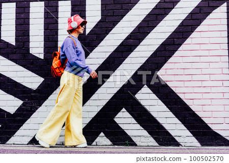 Side view stylish woman in bright clothes and bucket hat wearing wireless headphones and listening to music while walking near city urban painted wall. Fashionable hipster lifestyle. Selective focus. 100502570