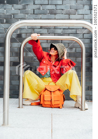 Hipster fashion young woman in bright clothes, sun glasses and bucket hat sitting near gray brick wall background with bike stands frame. Urban city street fashion. Vertical card. Selective focus. 100502571