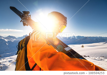 The skier stands on the top of the mountain and enjoys the view of the beautiful winter mountains on a sunny day. Man holding skis on his shoulder The skier stands on the top of the mountain and enjoys the view of the beautiful winter mountains on a sunny day. Man holding skis on his shoulder 100504253