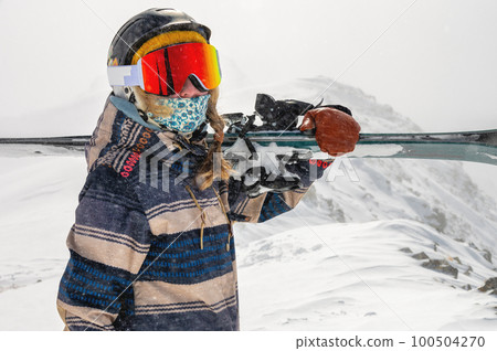 girl in a jacket carries skis on her shoulder against the backdrop of mountains at a ski resort. Image of a woman with skis on her shoulder against the backdrop of a snowy hill 100504270