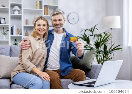 Portrait of happy family at home, adult mature couple man and woman sitting on sofa in living room and looking cheerfully at camera, man holding bank credit card in hands, happy with online shopping. 100504624