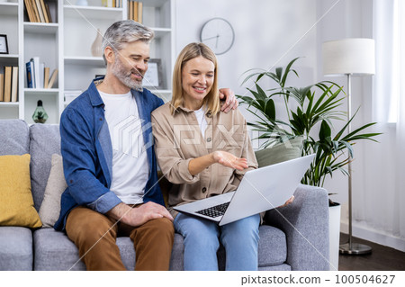 Senior mature couple man and woman sitting at home in living room on sofa, people in love using laptop, having fun choosing products in online internet store together. 100504627