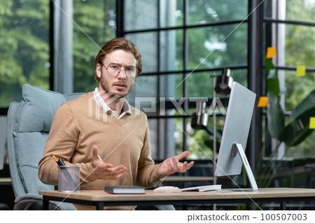 Solving a problem, making a decision. Portrait of a young freelancer, a programmer who looks worriedly and angrily at the camera, spreads his hands. Sitting in the office at the desk. 100507003