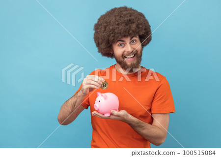 Portrait of man with Afro hairstyle wearing orange T-shirt investing in bitcoins, putting golden crypto coin into piggy bank, happy with money savings. Indoor studio shot isolated on blue background. Portrait of man with Afro hairstyle wearing orange T-shirt investing in bitcoins, putting golden crypto coin into piggy bank, happy with money savings. Indoor studio shot isolated on blue background. 100515084