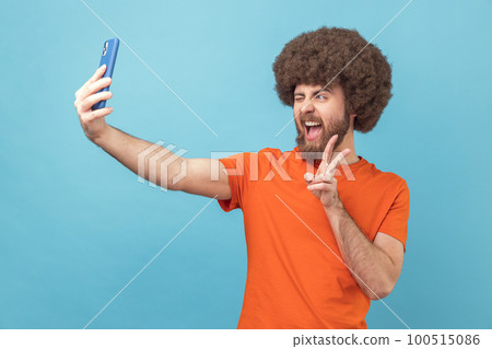 Portrait of man with Afro hairstyle wearing orange T-shirt showing v gesture, posing at phone camera, making selfie or having video call. Indoor studio shot isolated on blue background. Portrait of man with Afro hairstyle wearing orange T-shirt showing v gesture, posing at phone camera, making selfie or having video call. Indoor studio shot isolated on blue background. 100515086