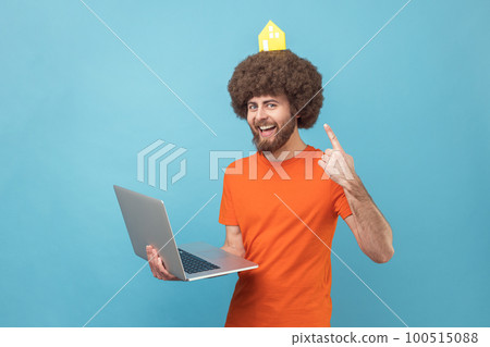 Portrait of funny man with Afro hairstyle wearing orange T-shirt holding notebook and pointing at paper house on his head, real estate. Indoor studio shot isolated on blue background. Portrait of funny man with Afro hairstyle wearing orange T-shirt holding notebook and pointing at paper house on his head, real estate. Indoor studio shot isolated on blue background. 100515088