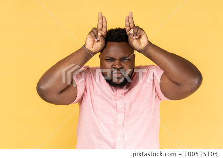 Portrait of man wearing pink shirt showing bull horn gesture, looking at camera with hostile angry expression, bully male threatening to attack. Indoor studio shot isolated on yellow background. 100515103