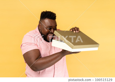 Portrait of optimistic man wearing pink shirt opening gift box and peeping inside with toothy smile, satisfied with present. Indoor studio shot isolated on yellow background. 100515129