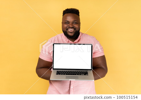 Portrait of happy smiling man wearing pink shirt showing laptop with blank display for advertisement, having satisfied expression. Indoor studio shot isolated on yellow background. 100515154