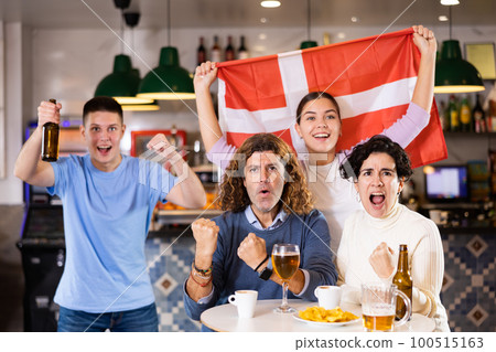 Company of young adult enthusiastic fans waving flag of Denmark and supporting national team with beer in pub 100515163