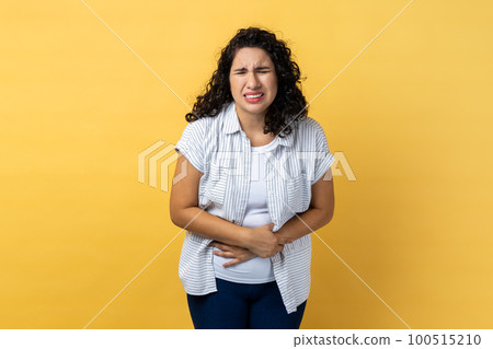 Portrait of sad depressed unhealthy woman with dark wavy hair suffering from strong stomachache, frowning, pain during period. Indoor studio shot isolated on yellow background. 100515210