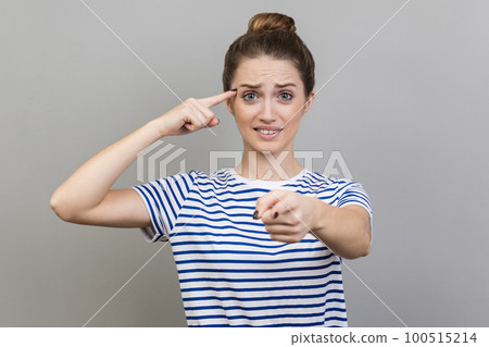 You are idiot. Woman wearing striped T-shirt showing stupid gesture and pointing to camera, blaming for insane plan, crazy idea, dumb suggestion. Indoor studio shot isolated on gray background. You are idiot. Woman wearing striped T-shirt showing stupid gesture and pointing to camera, blaming for insane plan, crazy idea, dumb suggestion. Indoor studio shot isolated on gray background. 100515214