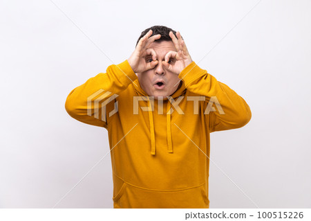 Portrait of curious man looks through fingers in binoculars gesture, observing distant with astonished look, watching afar, wearing urban style hoodie. Indoor studio shot isolated on white background. 100515226
