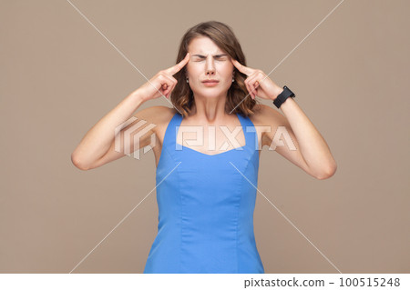 Sick unhealthy woman with wavy hair keeps fingers on temples, suffering terrible headache, standing with closed eyes, wearing blue dress. Indoor studio shot isolated on light brown background. 100515248