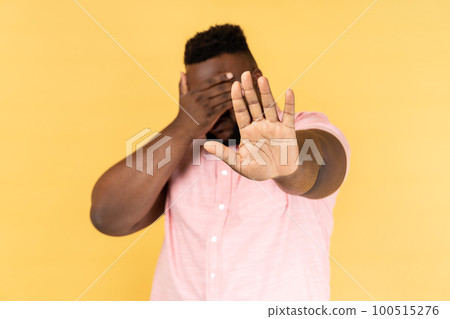 Stop, I don't want to see at this. Portrait of man wearing pink shirt covering his face and showing stop hand gesture. Indoor studio shot isolated on yellow background. Stop, I don't want to see at this. Portrait of man wearing pink shirt covering his face and showing stop hand gesture. Indoor studio shot isolated on yellow background. 100515276