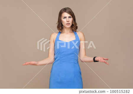 How could you. Annoyed indignant woman with wavy hair spreads palms, looks angrily at camera, quarrels with someone, wearing blue dress. Indoor studio shot isolated on light brown background. How could you. Annoyed indignant woman with wavy hair spreads palms, looks angrily at camera, quarrels with someone, wearing blue dress. Indoor studio shot isolated on light brown background. 100515282