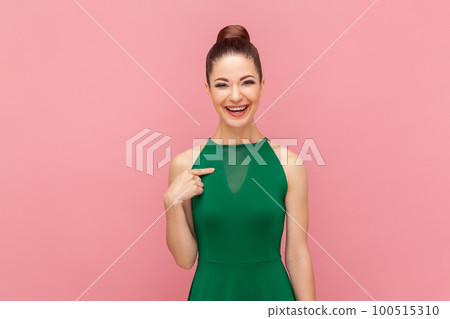 Portrait of smiling delighted attractive woman with bun hairstyle standing pointing at herself, being glad to be chosen, wearing green dress. Indoor studio shot isolated on pink background. 100515310