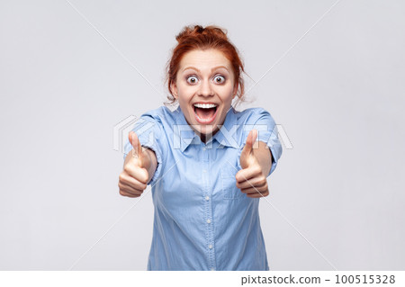 Portrait of positive redhead woman wearing blue shirt showing thumbs up, looking at camera with open mouth, surprised with excellent result. Indoor studio shot isolated on gray background. 100515328