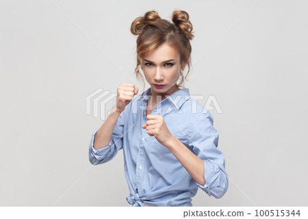 Woman having serious and confident look keeping fists clenched in front of her defencing herself from offence and bad treatment, wearing blue shirt. Indoor studio shot isolated on gray background. Woman having serious and confident look keeping fists clenched in front of her defencing herself from offence and bad treatment, wearing blue shirt. Indoor studio shot isolated on gray background. 100515344