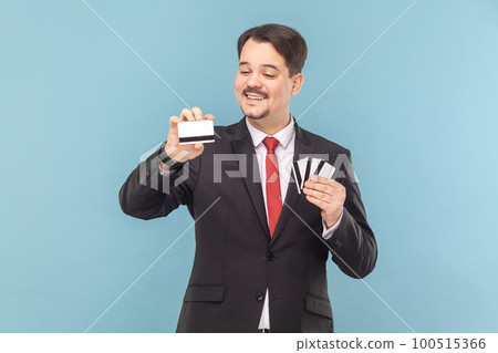 Portrait of delighted smiling greedy man with mustache standing holding many credit cards with big sum of money, wearing black suit with red tie. Indoor studio shot isolated on light blue background. 100515366