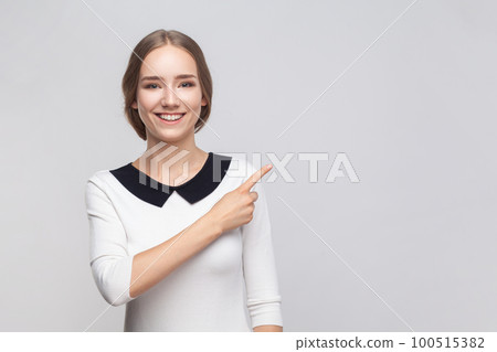 Portrait of pretty smiling optimistic woman wearing white dress pointing finger aside at advertisement area, copy space for promotion. Indoor studio shot isolated on gray background. Portrait of pretty smiling optimistic woman wearing white dress pointing finger aside at advertisement area, copy space for promotion. Indoor studio shot isolated on gray background. 100515382