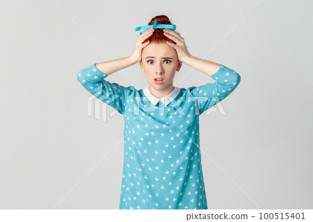 Portrait of astonished scared red haired woman with hair bun, standing with hands on her head, sees something shocked, wearing blue dress. Indoor studio shot isolated on gray background. Portrait of astonished scared red haired woman with hair bun, standing with hands on her head, sees something shocked, wearing blue dress. Indoor studio shot isolated on gray background. 100515401