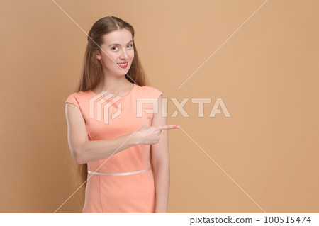 Portrait of smiling positive friendly woman with long hair pointing aside at advertisement area, copy space for promotion, wearing elegant dress. Indoor studio shot isolated on brown background. Portrait of smiling positive friendly woman with long hair pointing aside at advertisement area, copy space for promotion, wearing elegant dress. Indoor studio shot isolated on brown background. 100515474