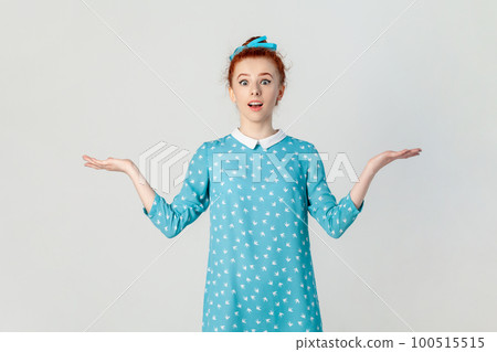 Portrait of shocked confused ginger woman standing with spread hands, looking at camera with big eyes, dosen't know how to react, wearing blue dress. Indoor studio shot isolated on gray background. Portrait of shocked confused ginger woman standing with spread hands, looking at camera with big eyes, dosen't know how to react, wearing blue dress. Indoor studio shot isolated on gray background. 100515515