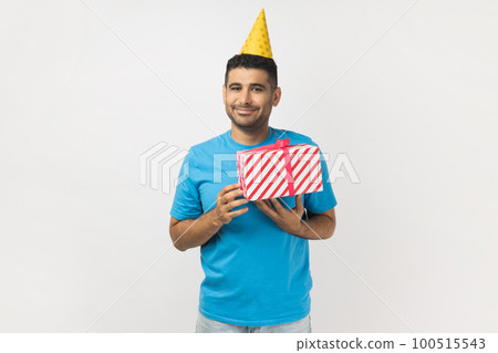 Portrait of festive smiling unshaven man wearing blue T- shirt and yellow party cone standing with present box, celebrating his birthday. Indoor studio shot isolated on gray background. 100515543