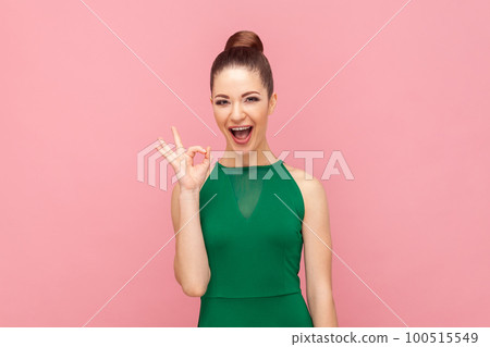 Portrait of excited amazed young adult woman showing ok sign, looking at camera with positive look, expressing happiness, wearing green dress. Indoor studio shot isolated on pink background. 100515549