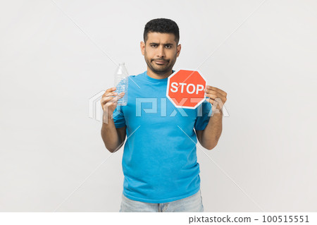 Portrait of sad serious unshaven man wearing blue T- shirt standing holding red stop symbol and empty plastic bottle, Earth protection. Indoor studio shot isolated on gray background. Portrait of sad serious unshaven man wearing blue T- shirt standing holding red stop symbol and empty plastic bottle, Earth protection. Indoor studio shot isolated on gray background. 100515551