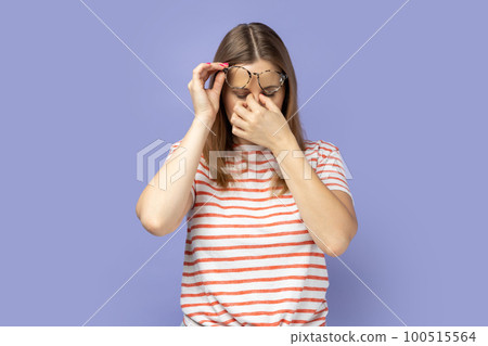 Portrait of frustrated blond woman wearing striped T-shirt touching closed eyes, crying from depression, grief or pain, feeling hopeless. Indoor studio shot isolated on purple background. 100515564