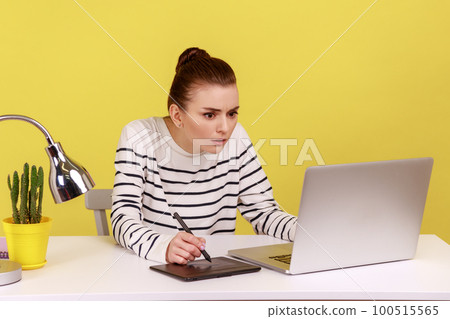 Concentrated busy serious woman working on graphic tablet, drawing sketches, sitting at workplace with laptop looking at display. Indoor studio studio shot isolated on yellow background. 100515565