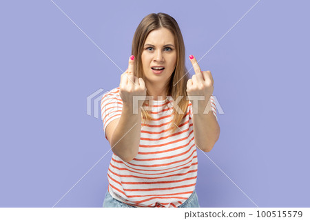 Portrait of rude vulgar blond woman wearing striped T-shirt showing middle fingers, impolite gesture of disrespect, looking with aggression hate. Indoor studio shot isolated on purple background. 100515579