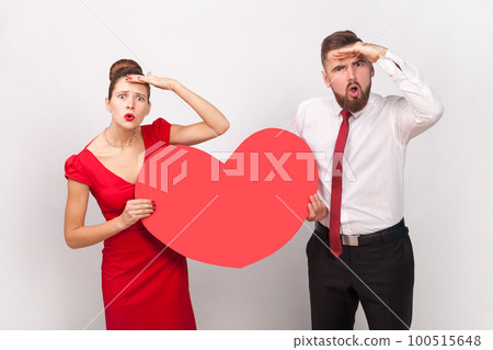 Portrait of surprised man in white shirt and woman in red dress standing together, holding big heart, looking far, waiting holiday. Indoor studio shot isolated on gray background. 100515648