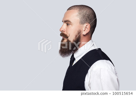 Side view portrait of serious attentive man with beard and mustache standing looking ahead with strict facial expression, being in bad mood. Indoor studio shot isolated on gray background. 100515654